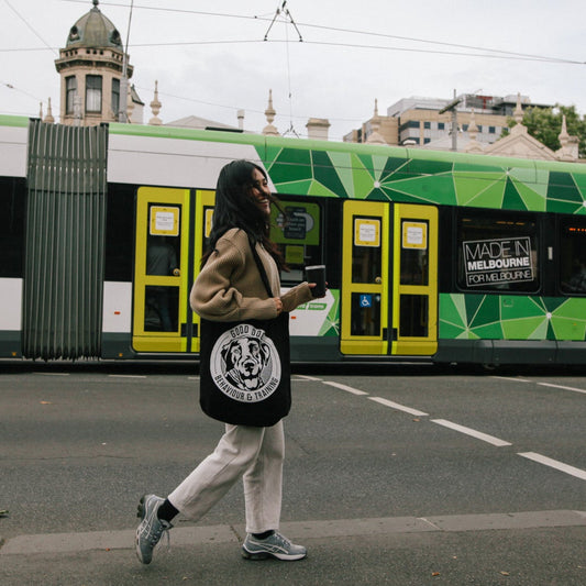 A woman walks across a street corner carrying a black tote bag with a white circular graphic, with a green and white modern tram in the background.