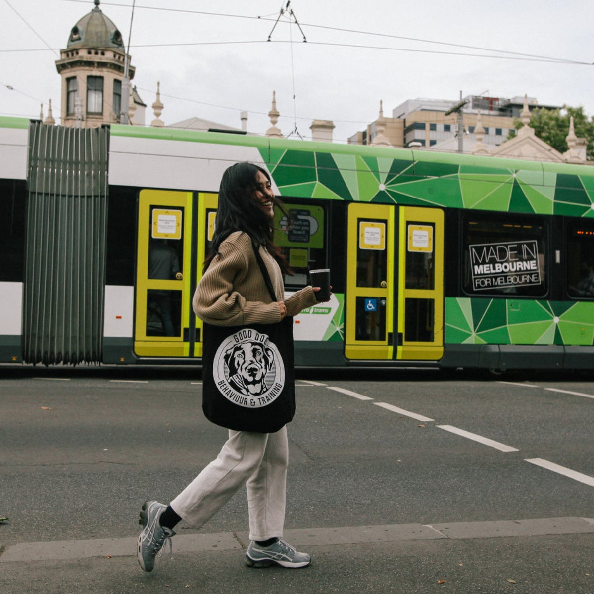 A woman walks across a street corner carrying a black tote bag with a white circular graphic, with a green and white modern tram in the background.