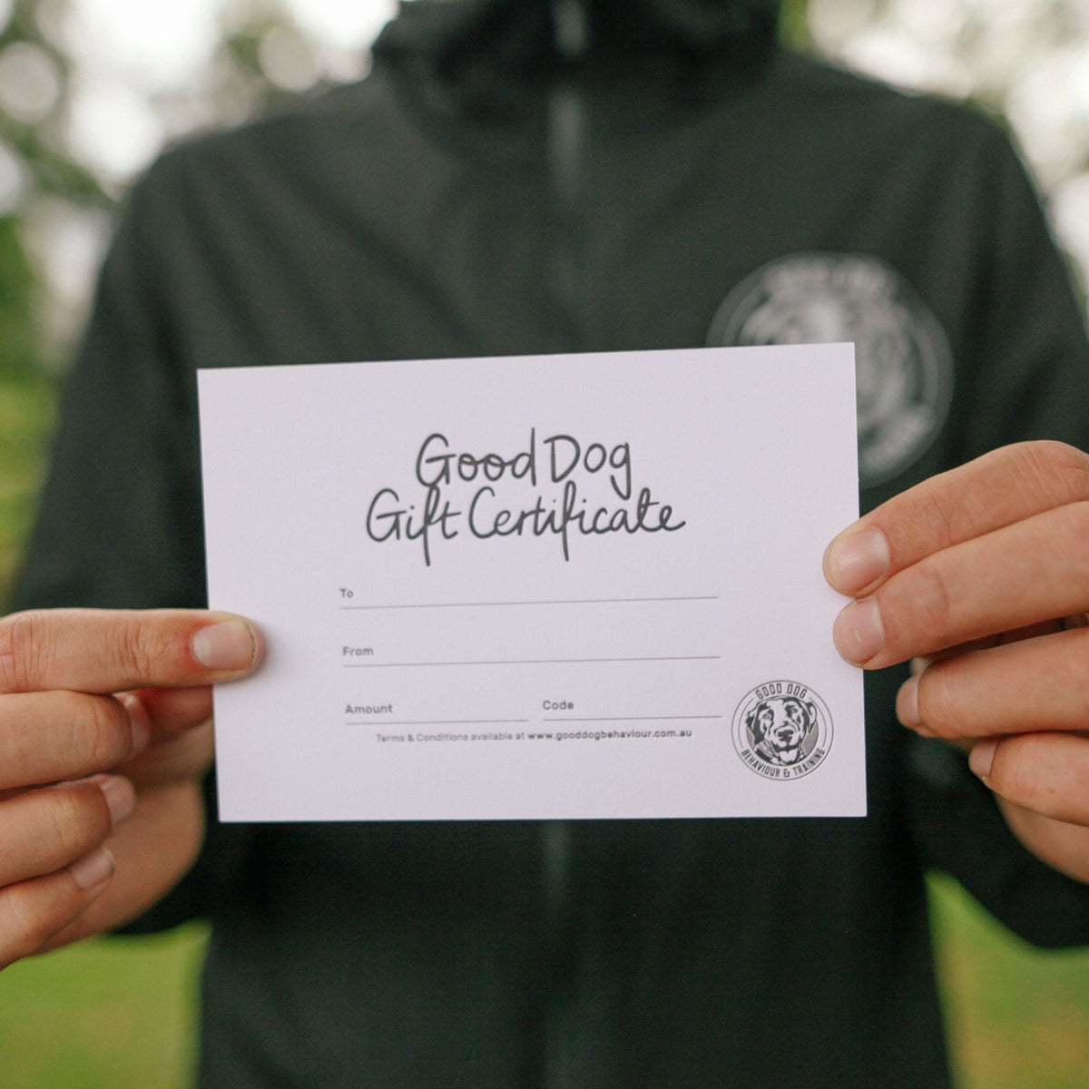 A person outdoors holds up a white gift certificate card with the text 'Good Dog Gift Certificate' written on it. The person is wearing a dark green hoodie, and the background is a blurred green outdoor setting.